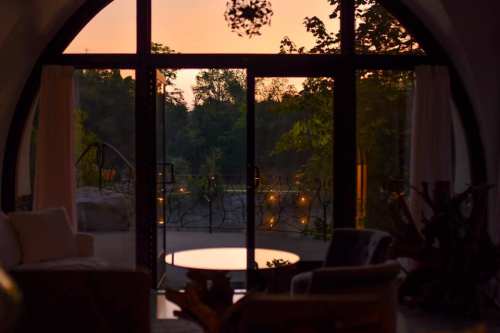 A cozy interior with a view of a sunset through large glass doors, surrounded by greenery and soft lighting.