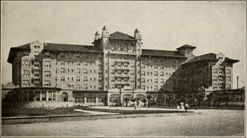 Historic hotel building with multiple stories, ornate architecture, and a large front lawn. Black and white photo.