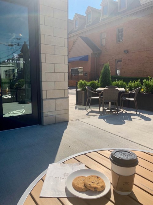 A sunny outdoor café scene with a table holding cookies and coffee, surrounded by greenery and a building in the background.