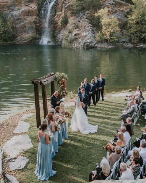 A wedding ceremony by a serene lake, with a waterfall in the background and guests seated on the grass.