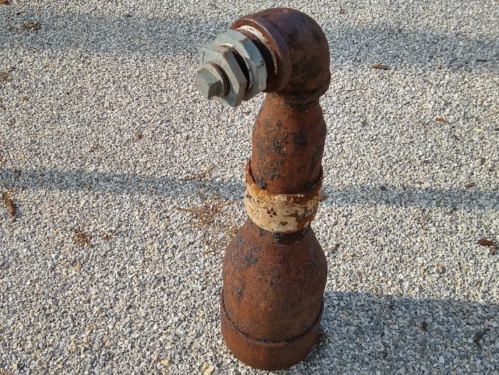 A rusty, curved metal pipe with a bolt on top, standing on a gravel surface.