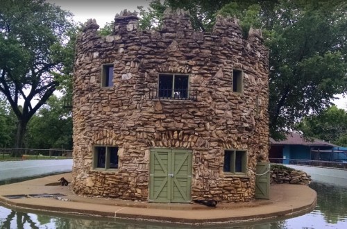 A stone castle-like structure surrounded by water, featuring windows and a green door, set in a park-like area.