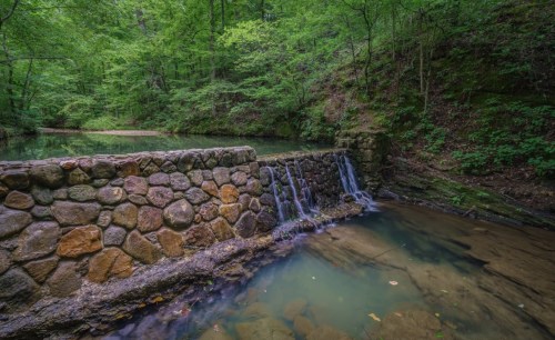 A serene stone wall with cascading water in a lush green forest by a calm stream.