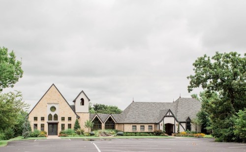 A charming brick church with a steep roof, surrounded by greenery and a cloudy sky. Parking lot in the foreground.