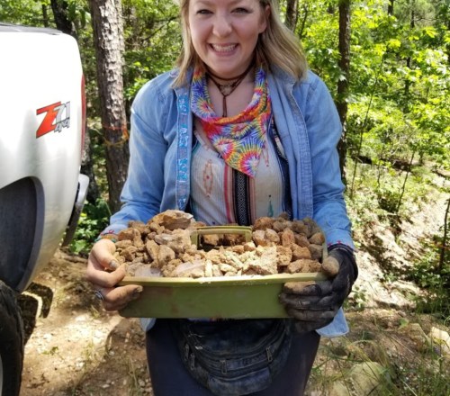 A smiling person holds a container filled with rocks, standing in a wooded area near a truck.