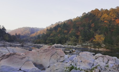 A serene river flows through rocky terrain, surrounded by trees with autumn foliage under a clear sky.