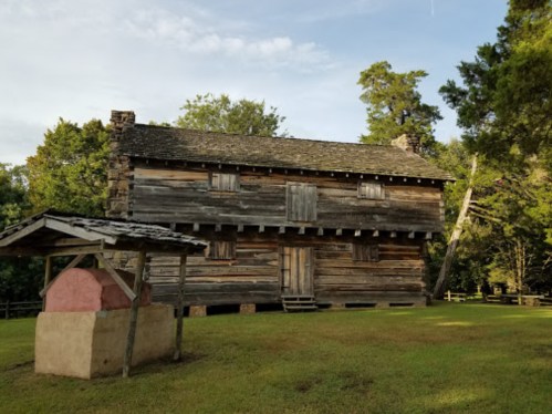 Historic wooden house with a stone well in front, surrounded by trees and grassy area.