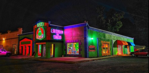 A colorful restaurant exterior at night, featuring neon lights and signs for ice cream and tacos.