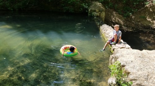 A child floats in a colorful inner tube in a clear pond while another child sits on the edge of the stone bank.
