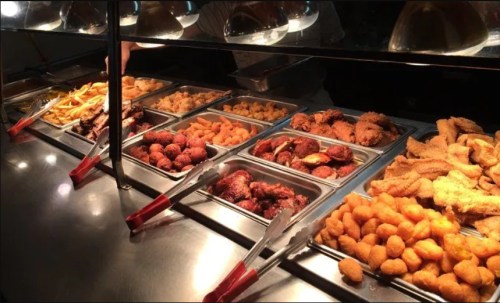 A buffet display featuring various fried foods, including chicken, fish, and sides, with serving tongs.