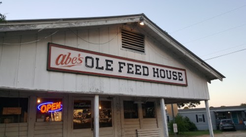Sign for Abe's Ole Feed House, a rustic building with an "Open" neon sign, during twilight.