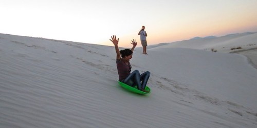 A person joyfully sleds down a white sand dune at sunset, with another person in the background taking a photo.