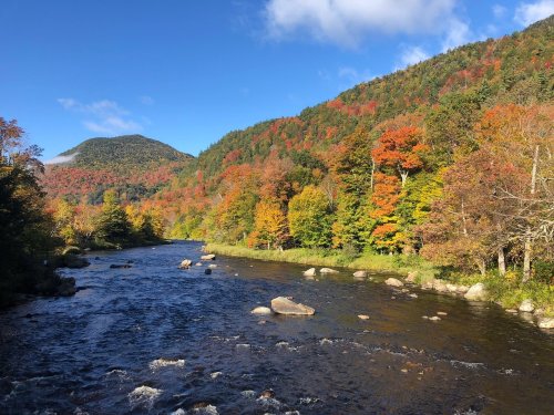 A serene river flows through a vibrant autumn landscape with colorful trees and mountains under a clear blue sky.