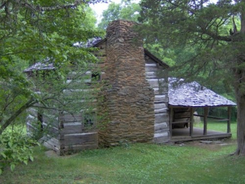 A rustic wooden cabin with a stone chimney, surrounded by greenery and trees.