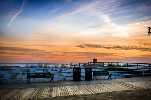 A serene beach boardwalk at sunset, featuring benches, trash cans, and a colorful sky with clouds.