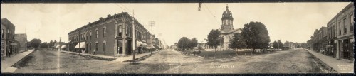 Historic panoramic view of a street in a small town, featuring buildings, trees, and a church in the background.