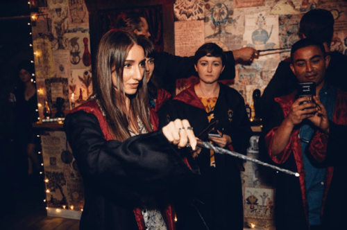 A group of people in wizard robes, one casting a spell with a wand, in a decorated, dimly lit room.