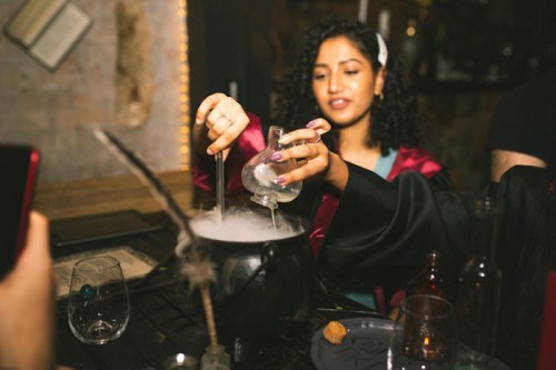 A person in a robe pours liquid from a glass into a cauldron, creating a misty effect in a dimly lit setting.
