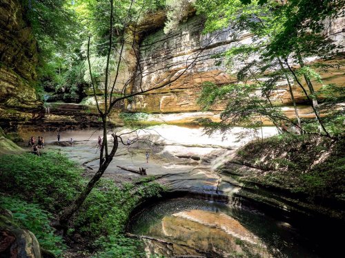 A serene natural landscape featuring lush greenery, rock formations, and a calm stream with people exploring the area.