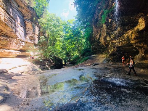 A scenic canyon with lush greenery, sunlight filtering through trees, and people exploring near a waterfall.