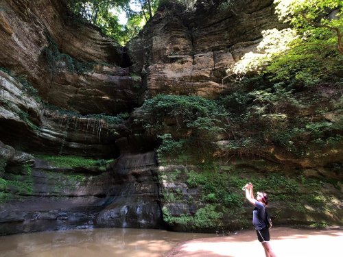 A person stands by a serene river, taking a photo of towering rock formations and lush greenery above.