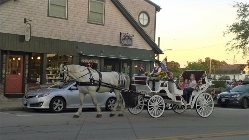 A white horse pulls a carriage with passengers, passing by a quaint shop at sunset.