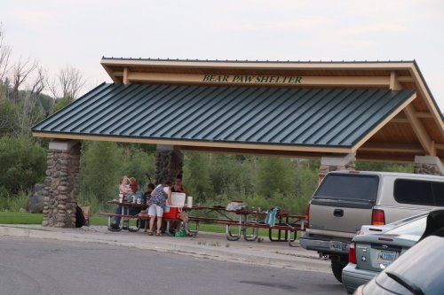 A group of people gathers under the Bear Paw Shelter, with picnic tables and a vehicle parked nearby.