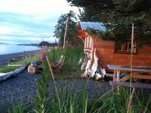 A cozy wooden cabin by the beach, surrounded by greenery and wildflowers, with a calm ocean in the background.
