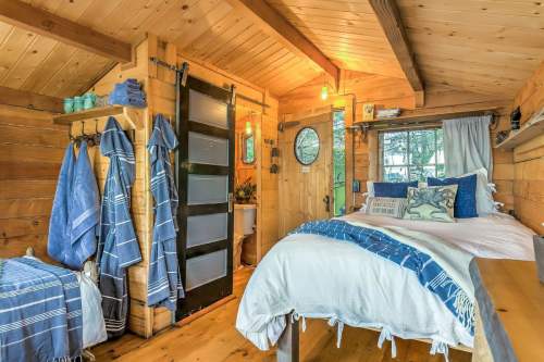 Cozy wooden interior of a small cabin with a bed, blue accents, and a sliding door leading to a bathroom area.