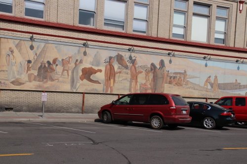 A mural depicting Indigenous people and scenes of daily life, set against a brick building, with parked cars in front.