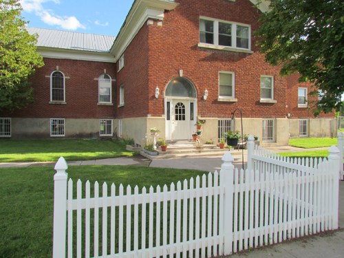 A brick building with large windows, a white door, and a picket fence, surrounded by green grass and trees.