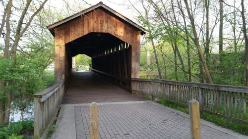 A wooden covered bridge leads into a lush green forest, with a paved path and wooden railings.