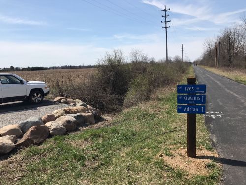 A gravel parking area beside a paved path, with directional signs for Ives, Kiwanis, and Adrian, and fields in the background.