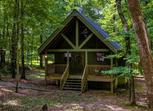 A green A-frame cabin surrounded by trees, featuring a front porch and steps leading to the entrance.