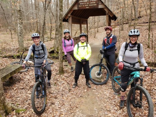 A group of five mountain bikers poses with their bikes near a wooden shelter in a forested area.