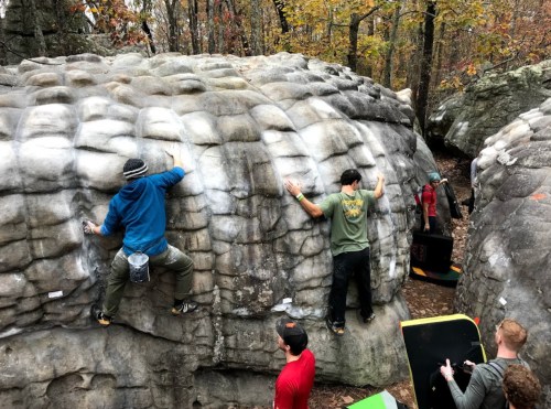 A group of climbers bouldering on large rock formations in a forested area during autumn.