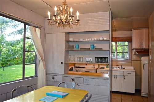 A vintage kitchen with a yellow table, shelves of dishes, and a chandelier, featuring large windows overlooking greenery.