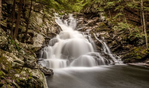 A serene waterfall cascades over rocky cliffs, surrounded by lush green trees and a calm pool below.