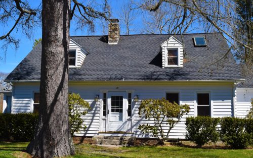 A white, two-story house with a gray roof, surrounded by greenery and a large tree in the front yard.