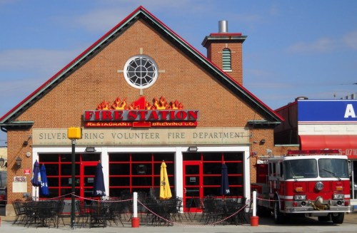 A brick building with a sign reading "Fire Station" and a fire truck parked outside, featuring outdoor seating.