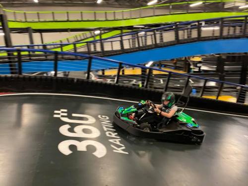 A person in a go-kart races around a curved track at a karting facility, with vibrant blue and green walls in the background.