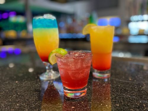 Three colorful cocktails on a bar counter: a layered blue, yellow, and orange drink, a red drink with ice, and a yellow drink.
