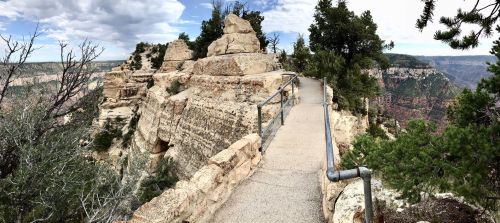 A scenic view of a rocky pathway along the edge of a canyon, surrounded by trees and dramatic cloud-filled skies.