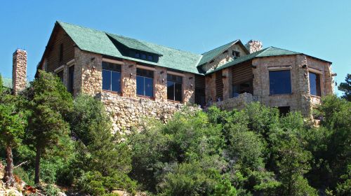A large stone and wood house with a green roof, surrounded by trees and set on a hillside under a clear blue sky.