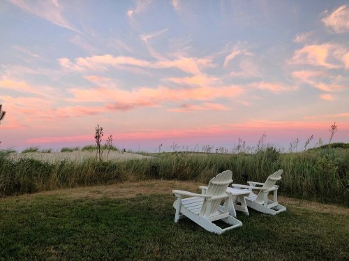 Two white Adirondack chairs sit on a grassy area, facing a colorful sunset over a serene landscape.
