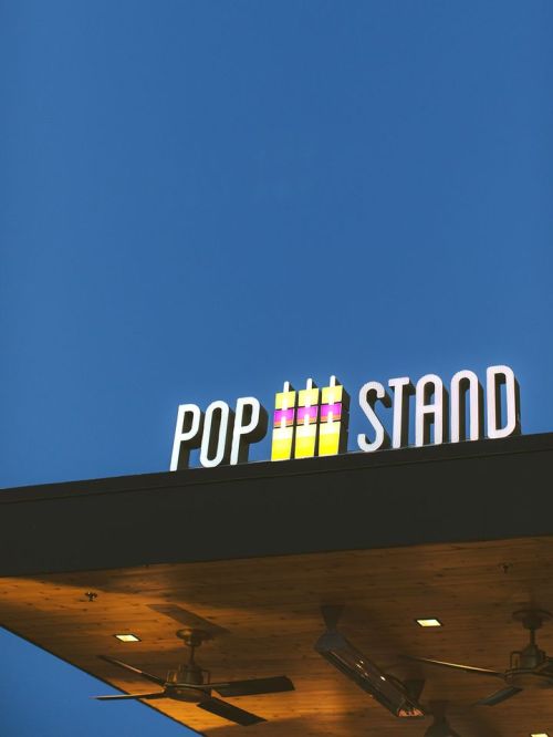 Sign for "POP STAND" illuminated against a clear blue sky, featuring colorful graphic elements.