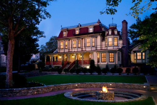 A large, elegant Victorian-style house at dusk, featuring a manicured lawn and a circular fire pit in the foreground.