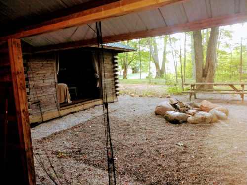 A rustic cabin with a fire pit surrounded by rocks, set in a wooded area with trees and a picnic table nearby.
