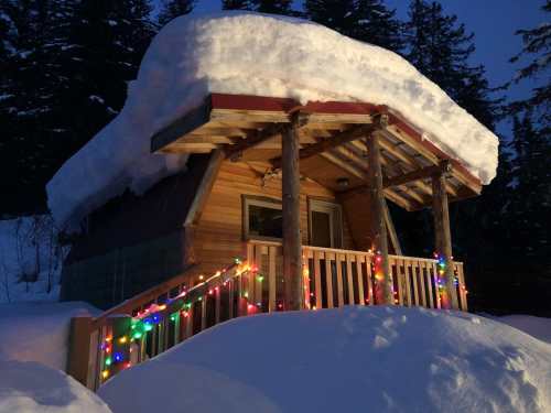 A cozy cabin covered in snow, adorned with colorful lights, surrounded by tall trees at dusk.