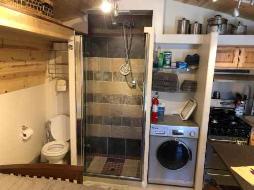 A compact bathroom with a shower, toilet, and laundry machine, adjacent to a small kitchen area in a cozy space.